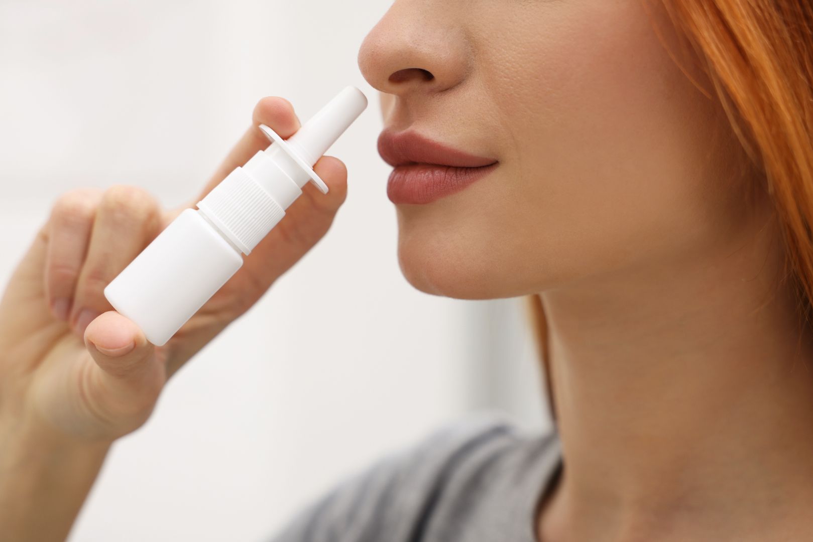 A close-up of a young woman with red hair about to use nasal spray. The spray is in a white container.