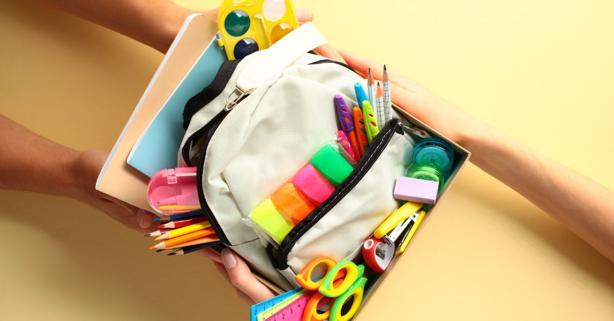 A close-up of someone donating a box of school supplies to a teacher. The box has a bookbag, writing utensils, and more.