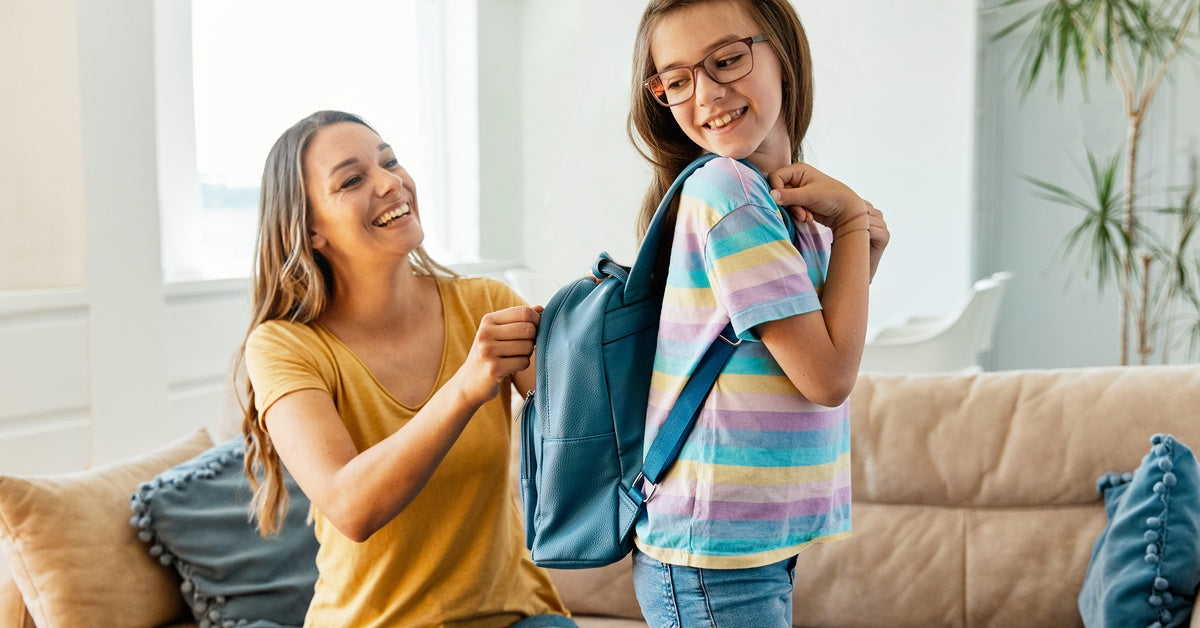 A woman sits on a couch and zips her daughter's backpack. The daughter smiles while looking over her shoulder at her backpack.