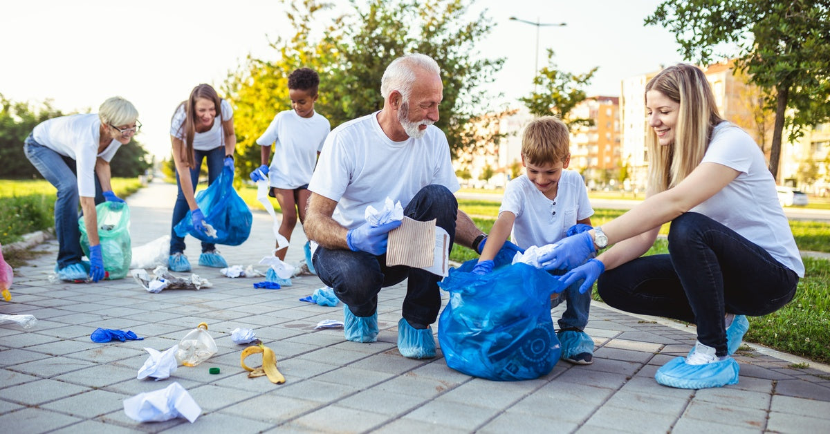A group of adults and children is picking up trash on a sidewalk. They all wear white T-shirts and blue gloves.