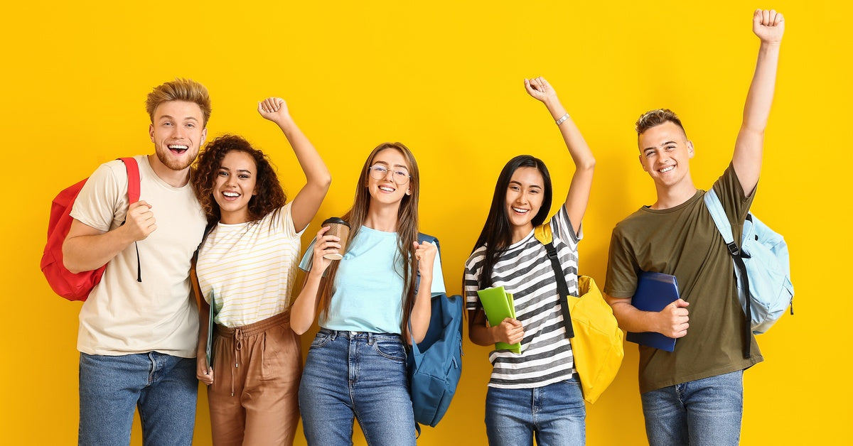 A group of smiling college students in front of a yellow background. The students wear backpacks and carry school supplies.