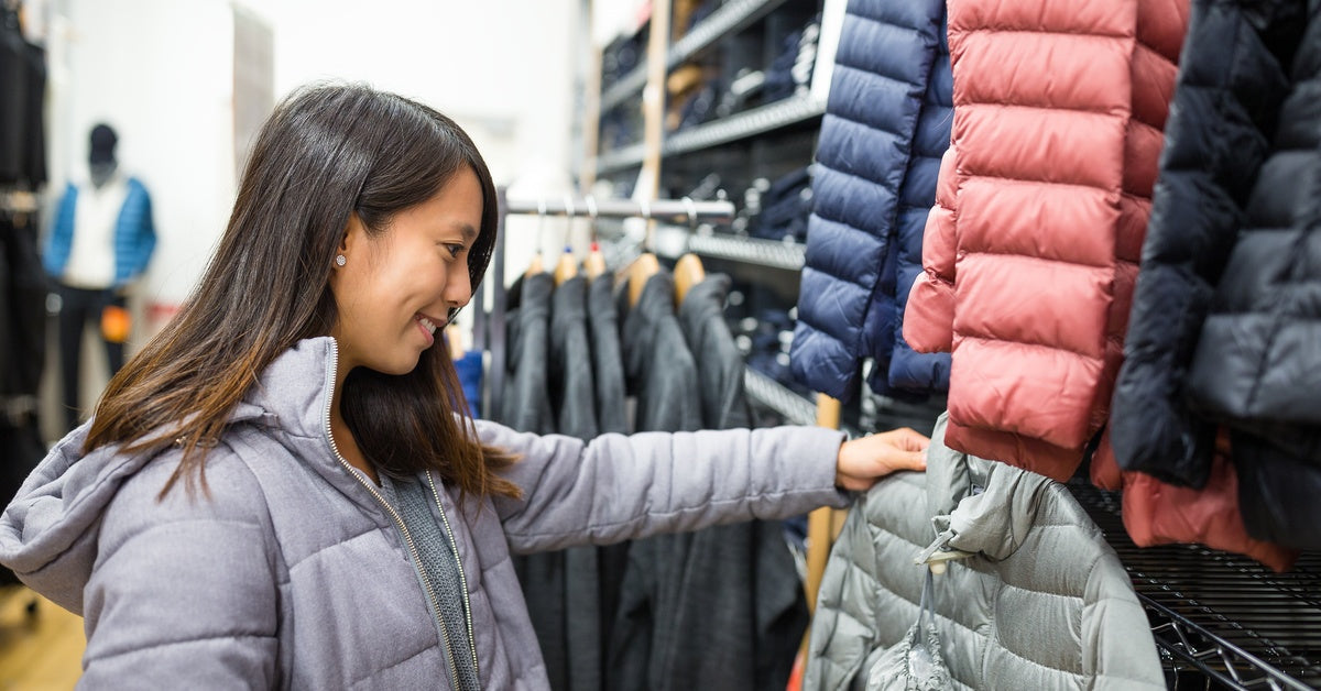 A woman holds a gray coat in front of a store coat rack. She's smiling, has brown hair, and wears a gray jacket.