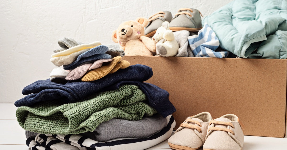 A box and a stack of children's clothing. The box also has a teddy bear and a pair of children's shoes.