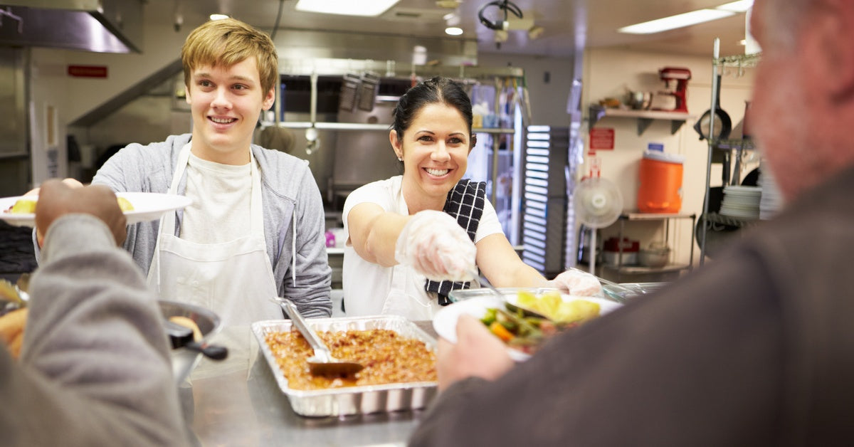 A man and a woman serve food in a homeless shelter. They both smile as they look toward a shelter resident.