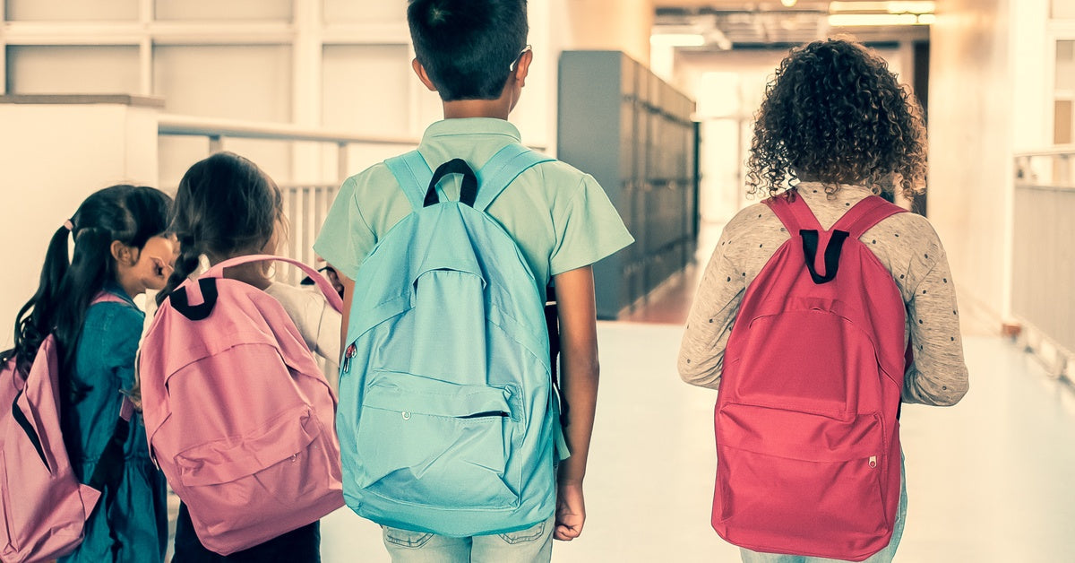 School-aged children walk together in a hallway with their backs facing the camera. They each wear a backpack.