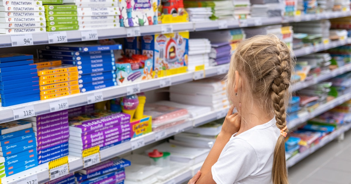 A young, blonde girl stands in front of a school supply shelf in a store. Her back faces the camera, and she holds her arm.