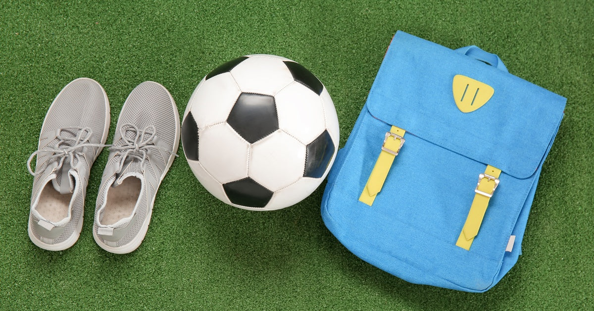 A pair of gray sneakers, a black-and-white soccer ball, and a blue backpack with yellow accents are lying on the grass.