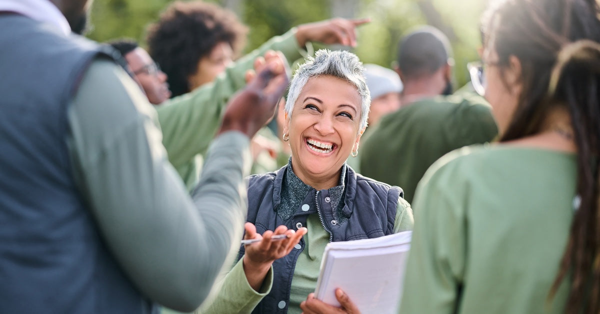 A woman with short grey hair is holding a notebook and smiling at a tall man while standing in a group of people who are all wearing green shirts.