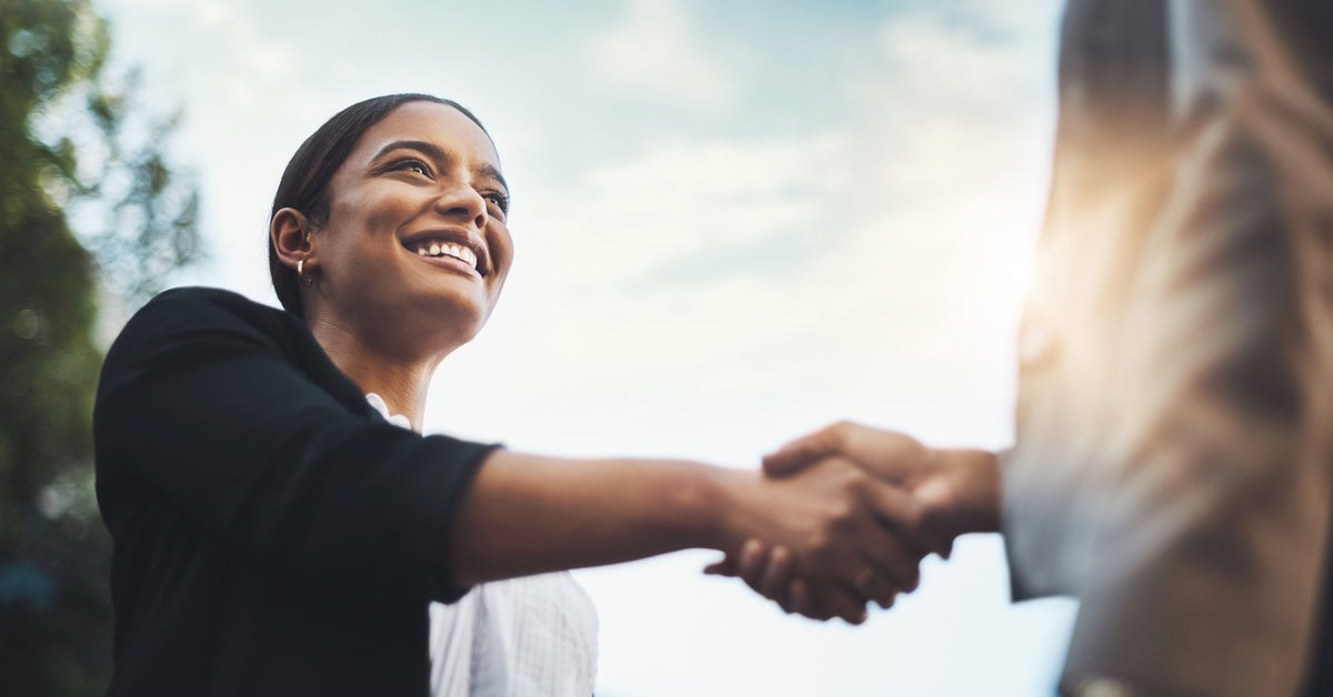 A smiling woman wearing a black jacket is greeting a person by shaking their hand.