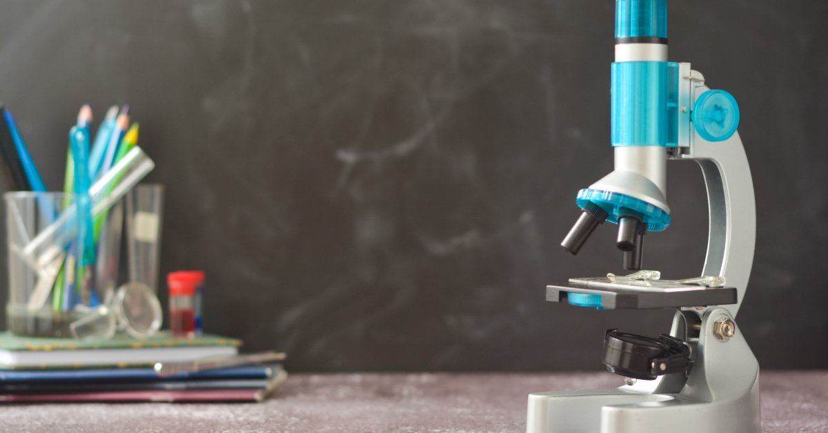 A microscope sits on a desk beside notebooks and pencils, set against a classroom chalkboard background.
