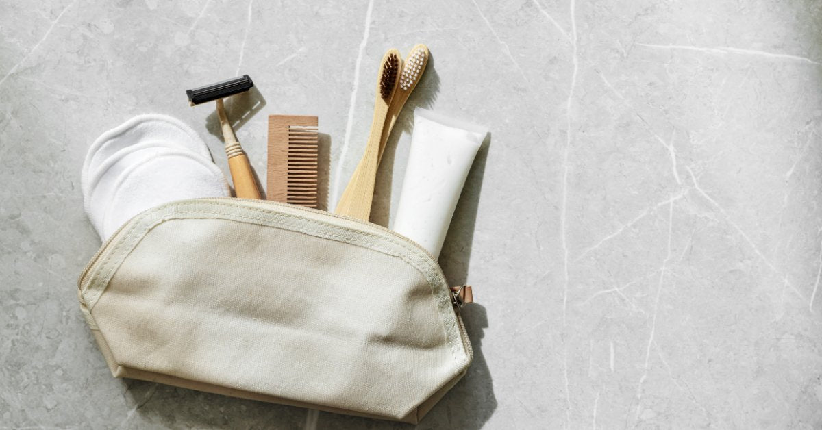 A toiletry bag holds bamboo toothbrushes, a wooden comb, a razor, and reusable pads on a gray surface.