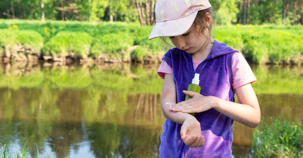 A young girl rubbing bug repellent spray into her arm. She wears a T-shirt and hat, and stands near a lake.