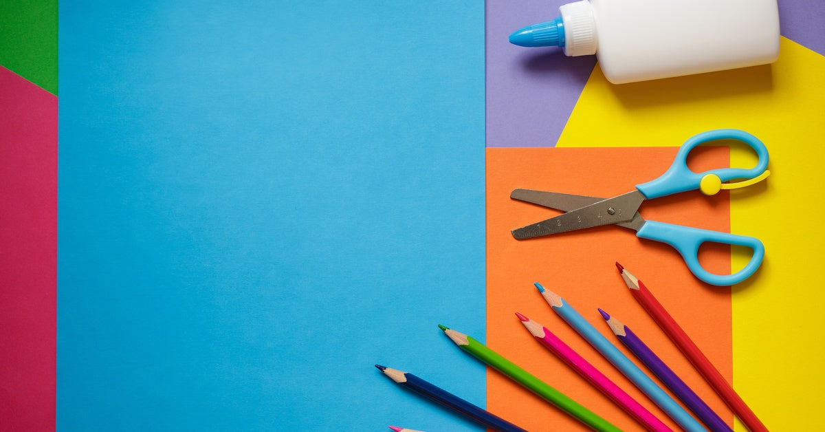 A bottle of school glue, a pair of kids' scissors, and seven colored pencils are laid out on top of colorful construction paper.