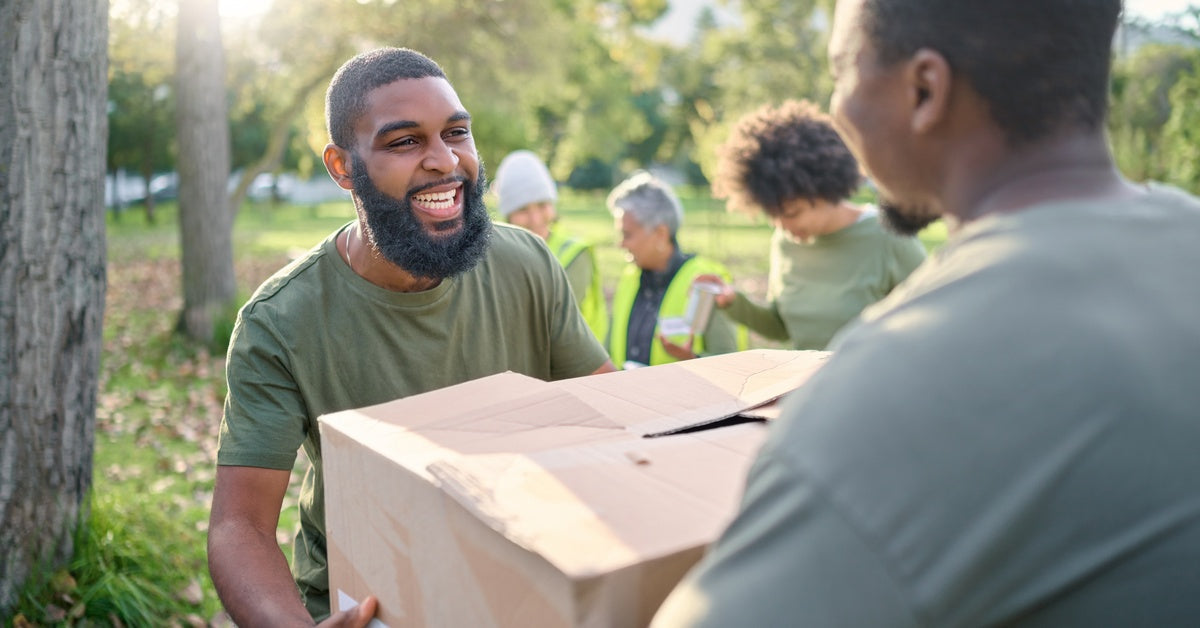 In a sunny park, a smiling community volunteer is giving a closed cardboard box to another man in a green shirt.