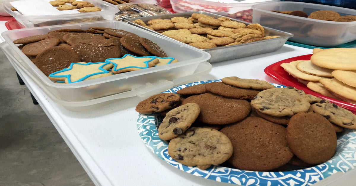 Plates and tupperware containers full of different types of cookies are placed on a white table for a bake sale event.