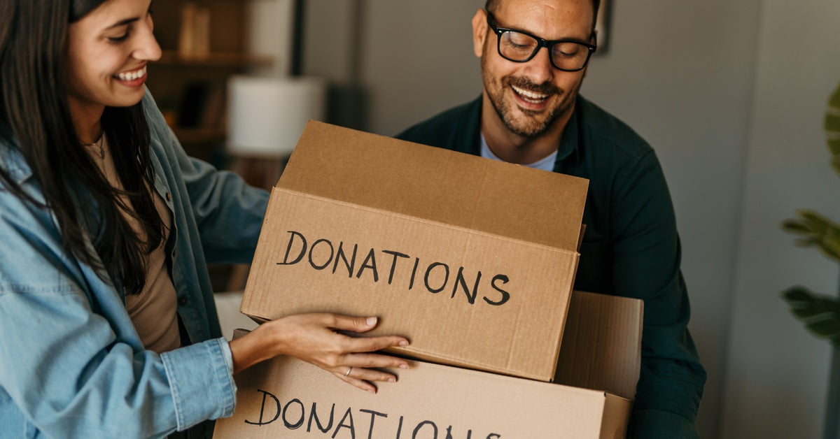 A smiling woman places two cardboard boxes with the word "DONATIONS" written on them into the arms of a smiling man.
