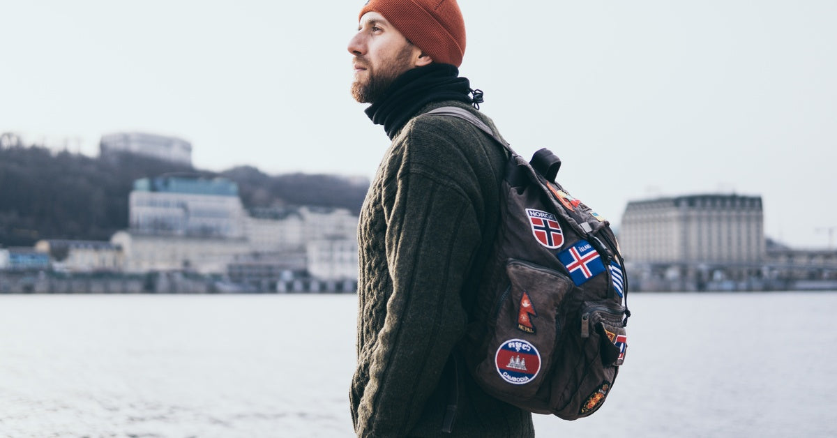 A bearded young man stands outdoors in the snow, wearing a sweater and an orange beanie, carrying a backpack with patches.
