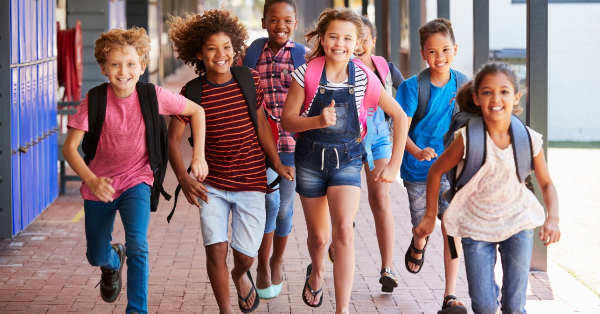 A group of smiling schoolchildren with backpacks run together down a hallway outside their classrooms.