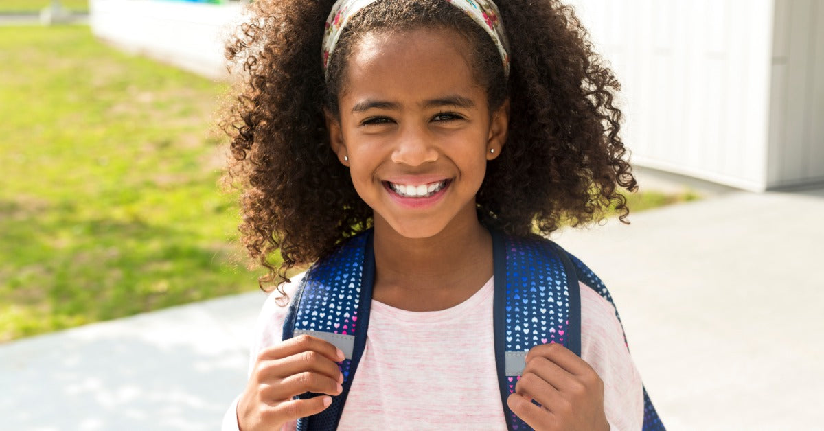 Smiling schoolgirl with curly hair wearing a backpack outdoors, standing on the sidewalk near the school building.