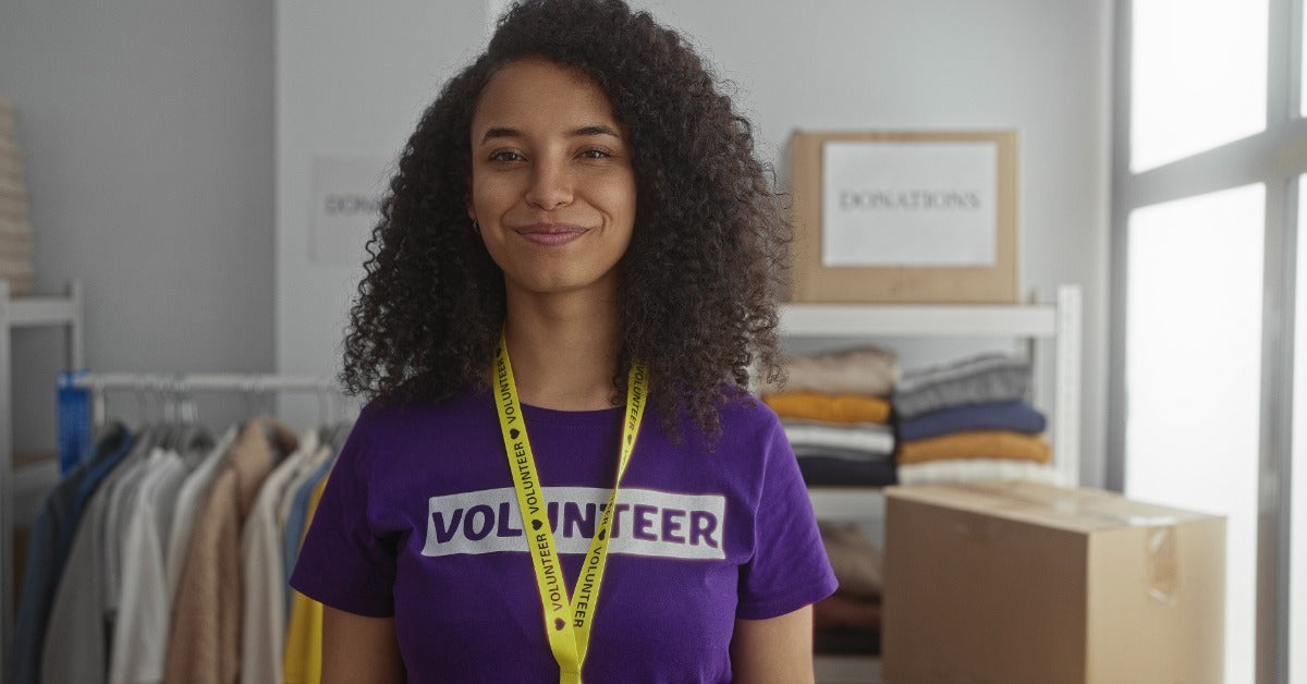 A smiling volunteer is wearing a purple shirt and stands in the donation center with folded clothes and boxes nearby.