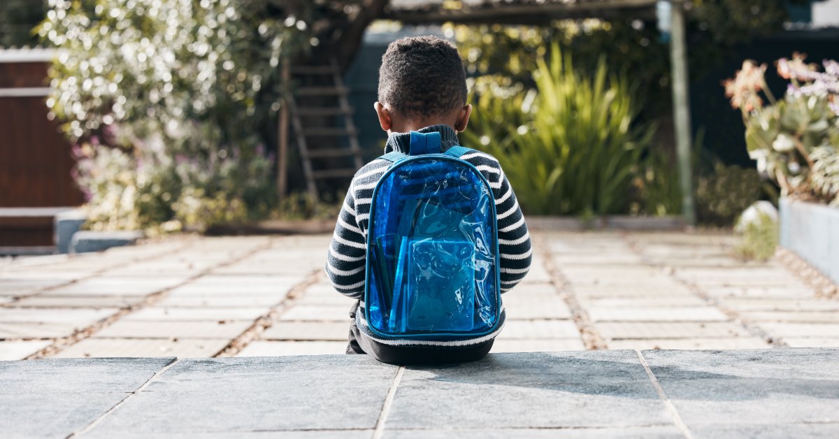 A young child is wearing a blue backpack and striped shirt while sitting on a patio in a garden on a sunny day.