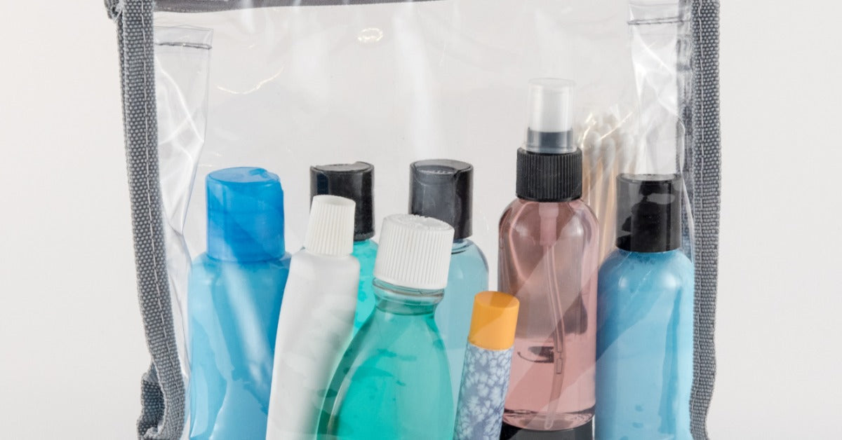A clear toiletry bag holds small bottles, a spray, and hygiene items arranged neatly against a white background.