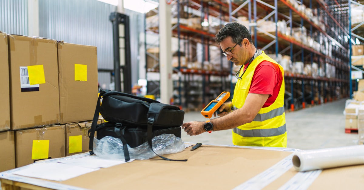 A warehouse worker scans a black backpack beside stacked shipping boxes in an organized storage facility.