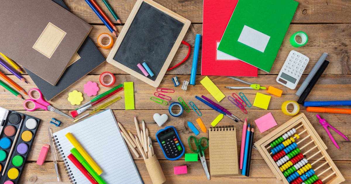 Assorted classroom supplies, including notebooks, pencils, scissors, and art materials, are arranged on a wooden table.
